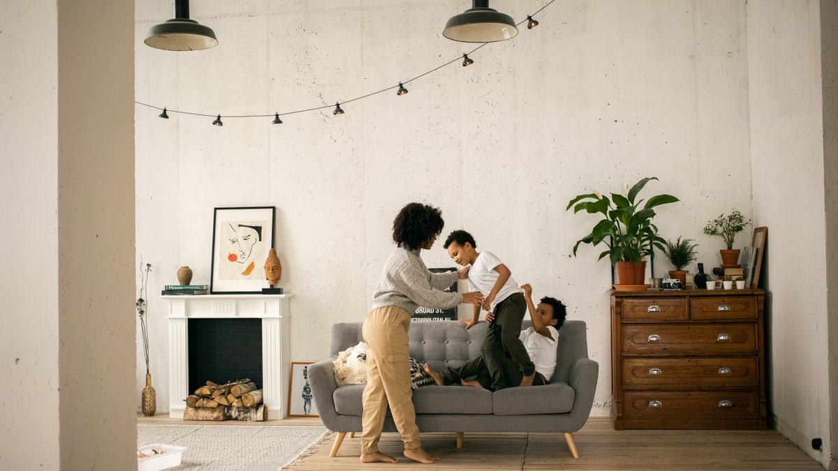 Full body of African American mother standing near sons having fun on sofa in light room with fireplace and hanging garland