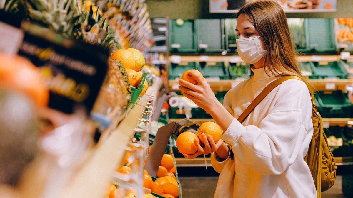 A woman wearing a face mask selects oranges in a grocery store, focusing on safety and prevention.